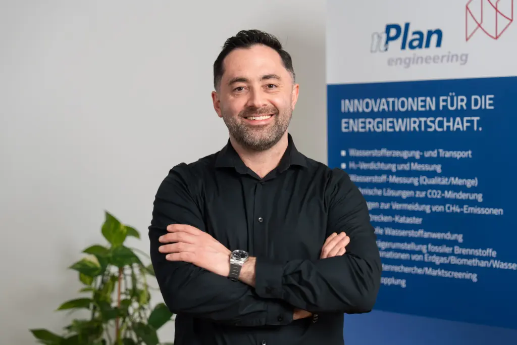 Ein lächelnder Mann in einem schwarzen Hemd steht mit verschränkten Armen in einem Büro in der Nähe einer Pflanze und eines blauen Technikbanners.