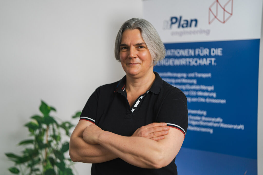 Eine Frau mit kurzen grauen Haaren steht mit verschränkten Armen lächelnd vor einem Firmenbanner und einer Pflanze.