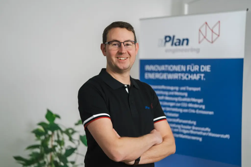 Ein lächelnder Mann mit Brille steht mit verschränkten Armen vor einem blau-weißen Ingenieurbanner.