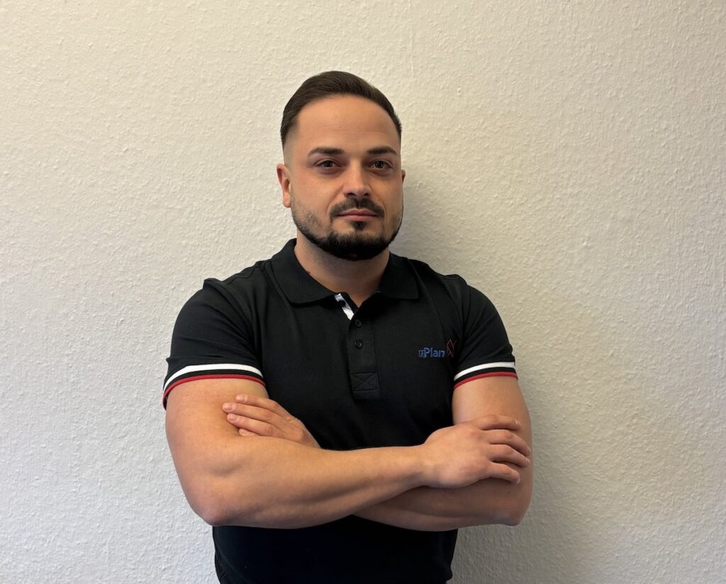 Ein Mann mit kurzen Haaren und Bart in einem schwarzen Polohemd steht mit verschränkten Armen vor einer schlichten Wand.